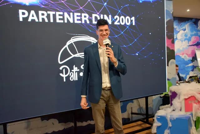 Un prezentator în sacou albastru vorbește la microfon în fața ecranului de parteneri.