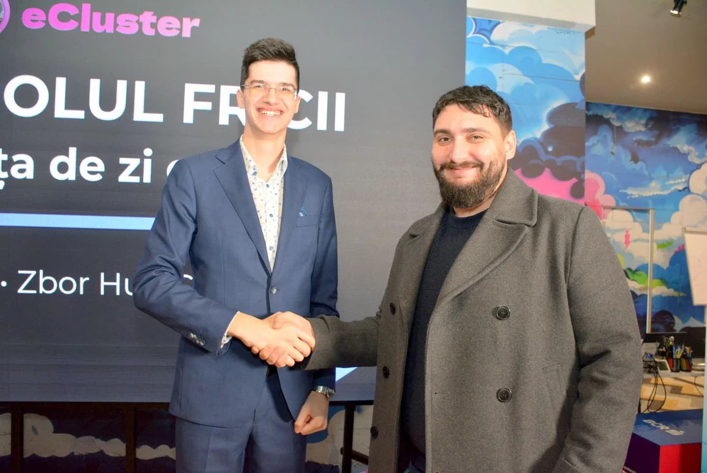Doi participanți zâmbesc și își strâng mâna în fața ecranului de eveniment.